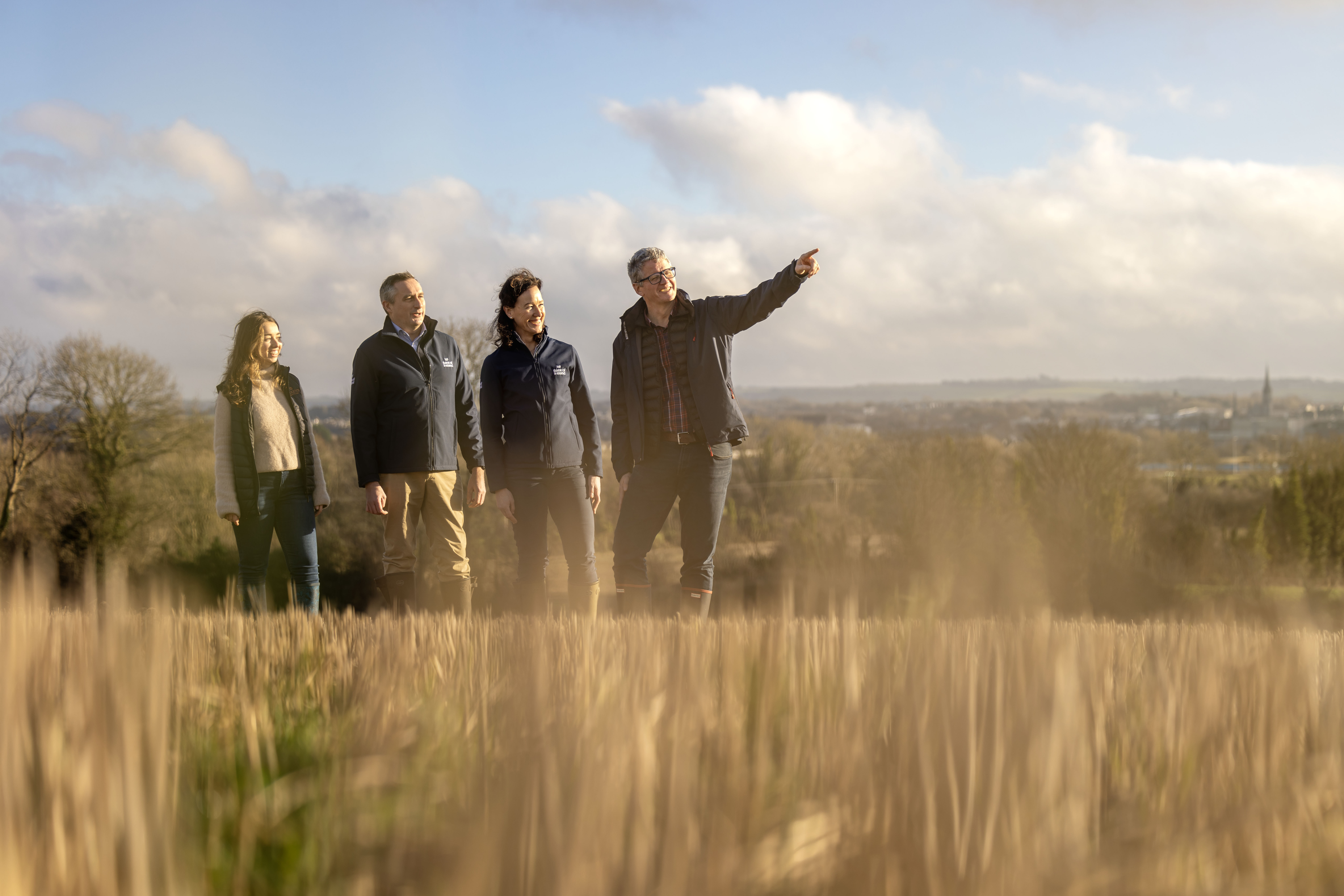Irish Distillers announces partnership with Bank of Ireland to offer Enviroflex loans to tillage farmers to incentivise farmers to continue implementing sustainability focused improvements on their land. The image shows four people standing in a field of barley on a sunny day with the man at the front of the group pointing into the distance.